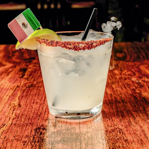 A smoking margarita with a chili-salt rim, lime, and a small Mexican flag on a wooden table.