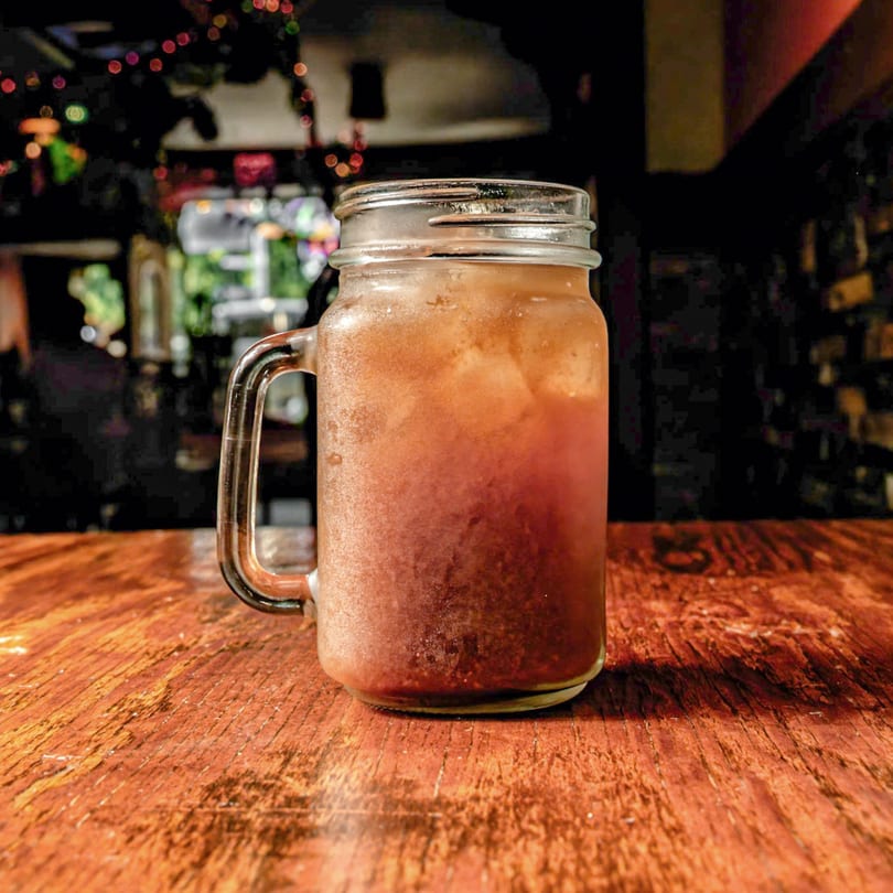 A frosty mason jar filled with a reddish-brown iced drink sits on a wooden table.