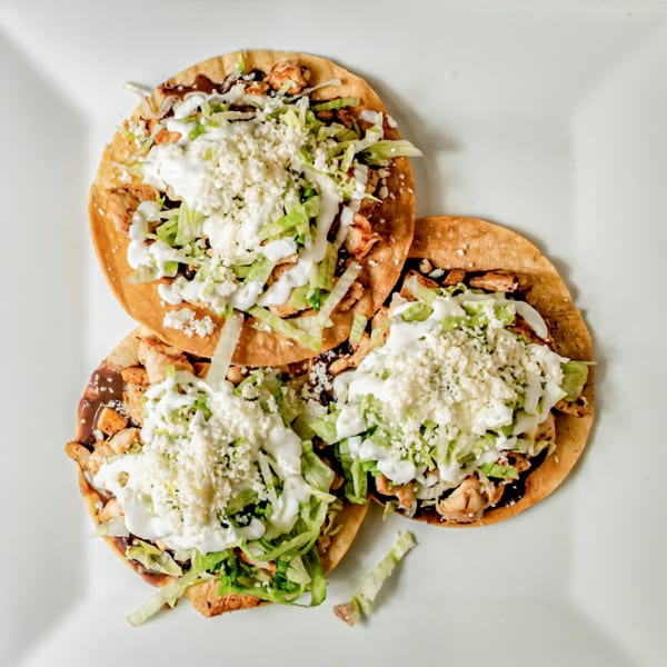 Three chicken tostadas with shredded lettuce, crumbled cheese, and crema on a white plate.