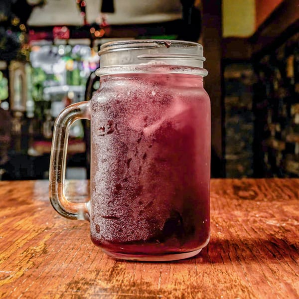 A frosty mason jar filled with a dark red beverage and ice sits on a wooden table.