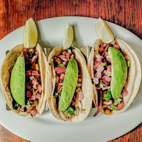 Three Avocados Mexican Grill tacos with meat, salsa, avocado slices, and lime wedges on a white plate.