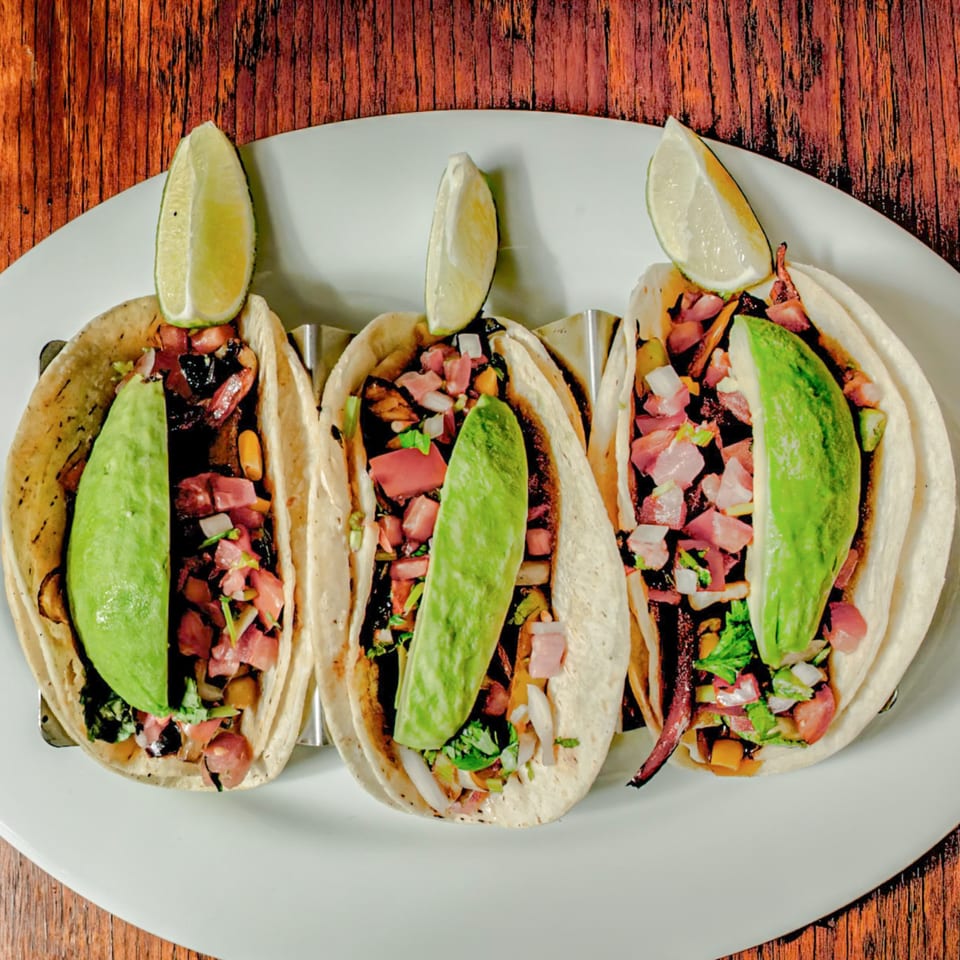 Three Avocados Mexican Grill tacos with meat, salsa, avocado slices, and lime wedges on a white plate.