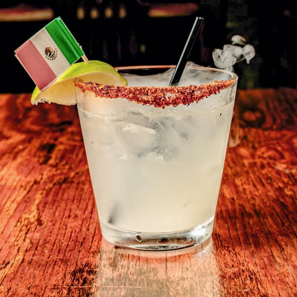 A smoking margarita with a chili-lime rim, lime wedge, and Mexican flag on a wooden table.
