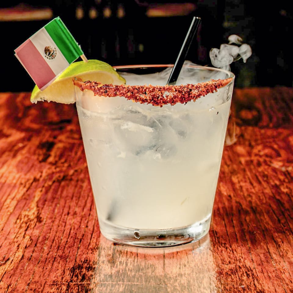 A smoking margarita with a chili-lime rim, lime wedge, and Mexican flag on a wooden table.