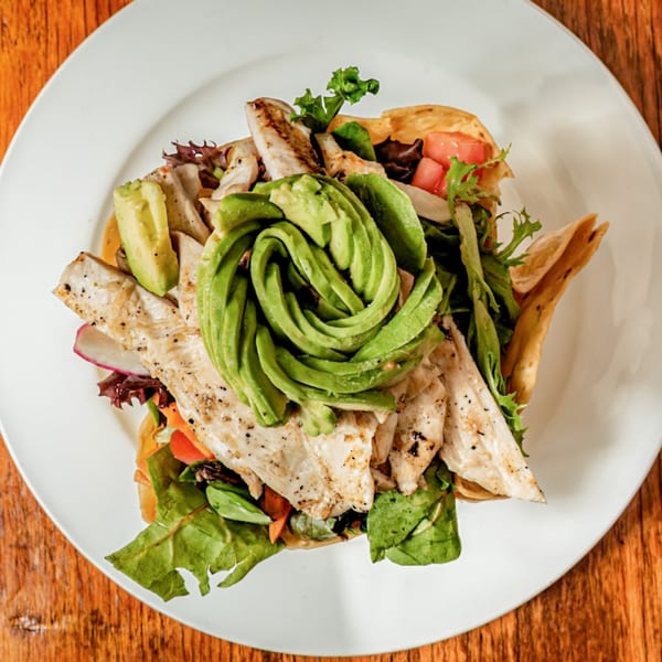 Grilled chicken salad with avocado, lettuce, and tomatoes in a tortilla bowl on a white plate.