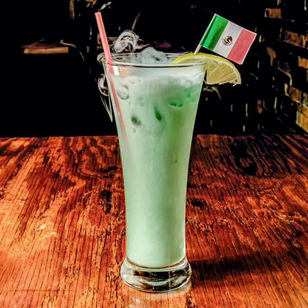 Tall glass of smoking green drink with a lime and Mexican flag on a wooden table.