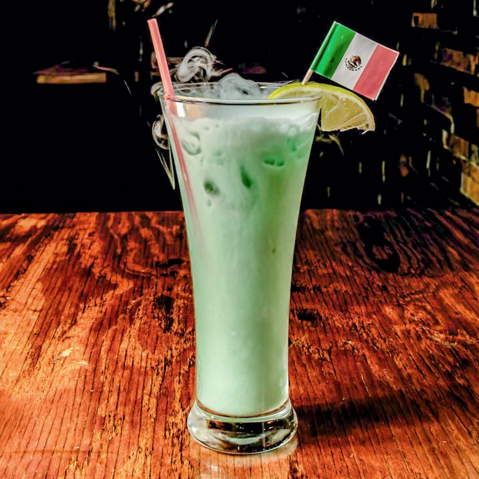 Tall glass of smoking green drink with a lime and Mexican flag on a wooden table.