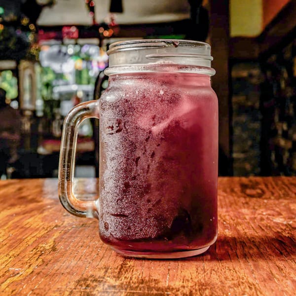 A frosty mason jar filled with a dark red drink and ice sits on a wooden table.