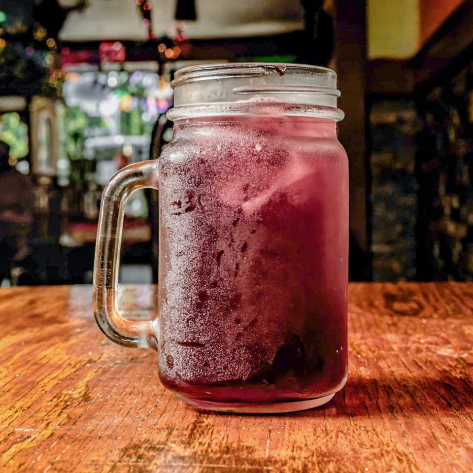 A frosty mason jar filled with a dark red drink and ice sits on a wooden table.