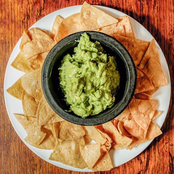 Avocados Mexican Grill's guacamole in a dark bowl, surrounded by tortilla chips on a white plate.