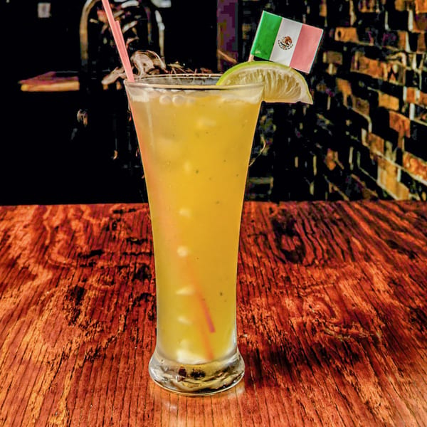 Tall glass of yellow drink with ice, lime, pink straw, and a small Mexican flag.