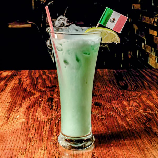 Smoking green cocktail with a Mexican flag and lime wedge on a rustic wooden table.