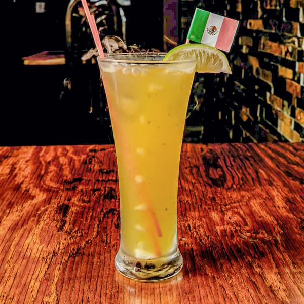 Tall glass of yellow drink with ice, lime, pink straw, and a Mexican flag on a wooden table.