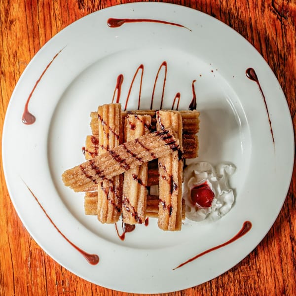 Churros with chocolate drizzle, whipped cream, and a cherry on a white plate.